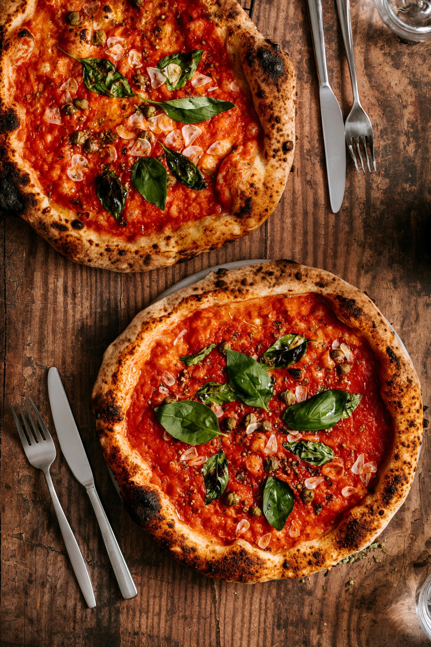Top view of two Neapolitan pizzas with fresh ingredients and silverware on a rustic wooden table.